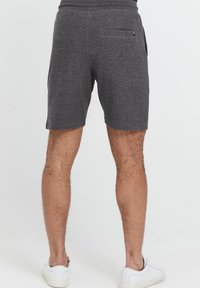 Graue Stoffshorts mit glatter Textur, ausgestattet mit einer Gesäßtasche und einer lockeren Passform. Kombiniert mit weißen Sneakers an nackten Beinen.