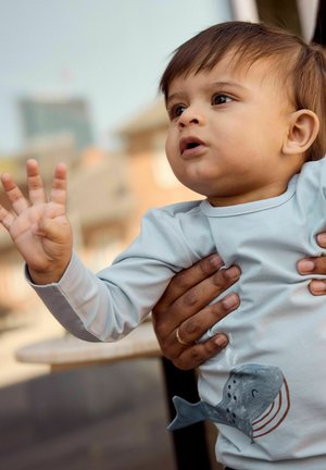 Niño pequeño con camiseta azul claro con diseño de ballena extendiendo la mano, sostenido de forma segura por manos adultas con un anillo dorado en un dedo.