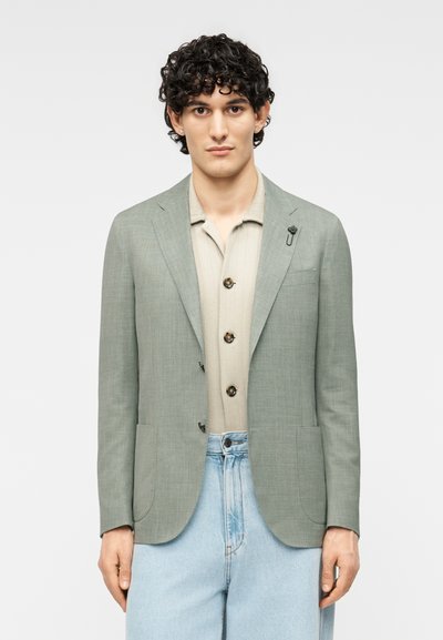 Blazer vert clair à finition texturée, avec des revers crantés, deux poches avant et une épingle florale sur le revers, associé à une chemise beige.