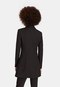 Persoon met krullend haar die een aansluitende lange zwarte blazer en zwarte broek draagt, staande met de rug naar de camera tegen een effen achtergrond.
