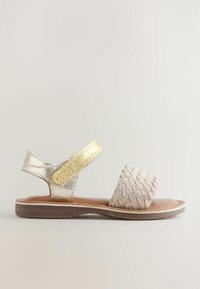 Dziecięca sandałka ze złotym paskiem wokół kostki i jasnobeżowym, plecionym paskiem z przodu na brązowej gumowej podeszwie, widok z boku na neutralnym tle.