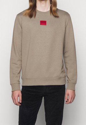 Beige Sweatshirt mit langen Ärmeln und rotem HUGO-Logo auf der Brust, getragen von einer Person mit schulterlangen Haaren und schwarzer Hose.