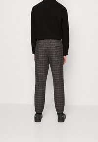 Schwarzes Hemd kombiniert mit karierten Hosen. Die Hosen zeigen ein kariertes Muster in dunklen und hellen Farben, mit subtiler Textur und schmal zulaufender Passform.