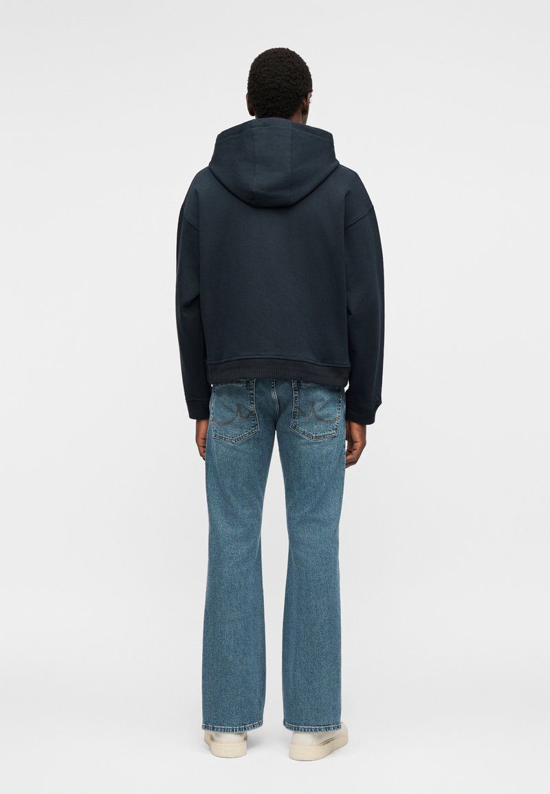 Homme portant un sweat à capuche noir, un jean bleu et des baskets blanches, debout de dos sur un fond uni.