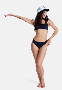 Ensemble bikini noir avec un haut ajusté et un bas taille haute. Porté avec une casquette de baseball blanche et noire. Le mannequin aux longs cheveux foncés pose.