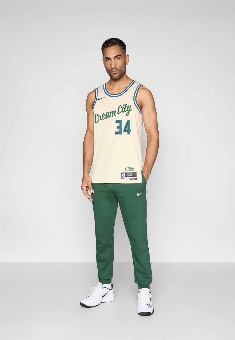 Maillot de basketball couleur crème avec "Cream City" et le numéro "34" en vert. Associé à un pantalon de survêtement vert et des chaussures de sport blanches.