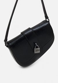 Sac à main en simili cuir noir avec une surface texturée, rabat pliant et quincaillerie argentée arborant une étiquette de verrou décorative. Bandoulière ajustable.