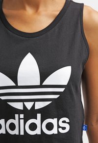 Närbild av en person som bär en svart Adidas linne med en stor vit trefoil-logotyp på bröstet.