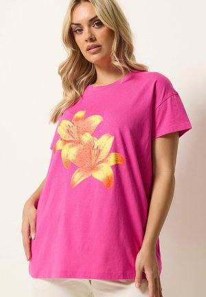 Blondynka w jasnoróżowej koszulce z krótkimi rękawami, z dużymi żółto-pomarańczowymi liliami nadrukowanymi z przodu, stojąca na jasnym tle.