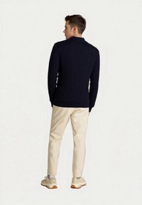 Mladý muž s krátkými hnědými vlasy, oblečený v tmavě modrém svetr s copánkovým vzorem, béžových kalhotách, bílých ponožkách a béžových teniskách, obrácený zády.