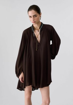 Donna che indossa un abito largo marrone scuro con maniche lunghe, scollo decorato e collane d'oro stratificate, in piedi contro uno sfondo chiaro e semplice.