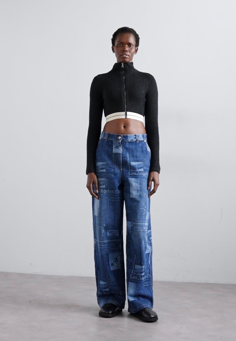 Haut zippé cropped en tricot noir, pantalon en denim bleu taille haute avec motifs imprimés, et chaussures noires chunky. Fond minimaliste.