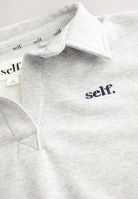 Hellgraues gefaltetes Poloshirt mit Kragen und schwarzem, besticktem Text "self." im Brustbereich.