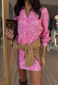 Γυναίκα που φοράει ροζ φόρεμα με κουμπιά και σχέδιο πασλέι, με καφέ πουλόβερ δεμένο γύρω από τη μέση, κρατώντας τσάντα φάκελο με σχέδιο λεοπάρ, στέκεται σε εσωτερικό χώρο.