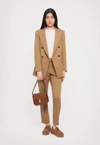 Brun skreddersydd dress med dobbelbrystet blazer og smale bukser. Modellen holder en brun crossover-veske og har på seg brune platåsko.