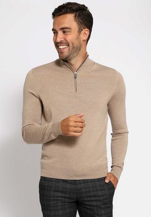 Man lacht, draagt een beige trui met rits en donkergrijze geruite broek, staat met één hand in de zak en de andere omhoog.