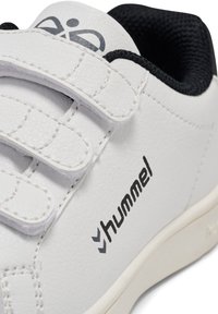 Vit lädersko med kardborreband, svarta accenter och texturerad yta. Har varumärkeslogotyp på sidan. Klassisk design, lämplig för fritidsbruk.