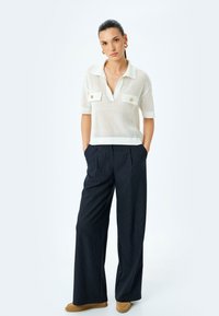 Polo bianco a maglia con maniche corte, dotato di due tasche sul petto e bottoni dorati. Abbinato a pantaloni a gamba larga blu con righe.