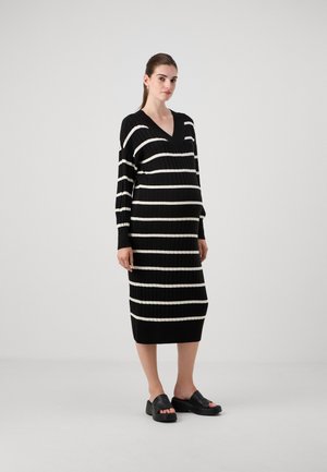 Zwart-witte gestreepte ribbenjurk met een V-hals, lange mouwen en een aansluitende silhouet, gecombineerd met zwarte slip-on schoenen met blokhak.