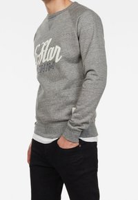 Sudadera de cuello redondo gris con estampado gráfico blanco y navy en la parte frontal. Hecha de tela texturizada, llevada sobre una camiseta blanca. Jeans negros.