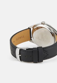 Montre pour homme avec un boîtier en acier inoxydable argenté, un bracelet en cuir noir et une boucle ardillon. Le dos présente un marquage en relief et des détails.