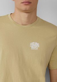 Beige katoen T-shirt met een ronde hals. Met op de linker borst het witte tekstlogo "EVOLVING ROOTS" en "ENDLESS GROWTH". Zachte textuur.
