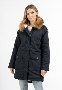 Parka imbottito blu con cappuccio bordato di pelliccia, due tasche frontali con dettaglio a bottone e chiusura con zip. Indossato sopra jeans chiari.