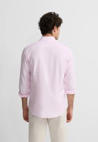 Camisa de manga larga rosa clara con las mangas remangadas, cuello abotonado y textura suave, llevada por una persona de espaldas.