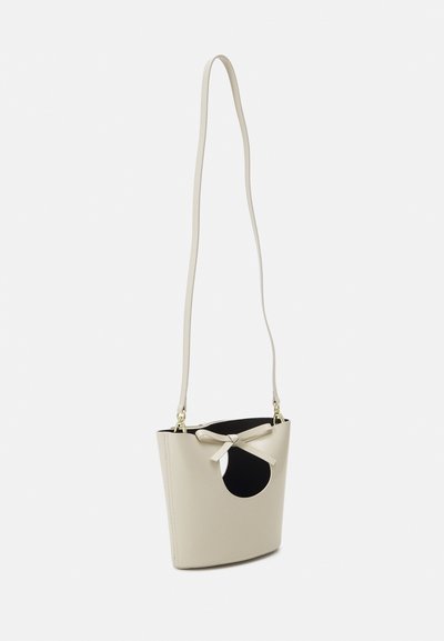 Small beige leather bucket bag with a circular cutout, bow detail on the front, and a long beige strap. Interior is black.
