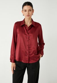 Blusa de satén burdeos con mangas largas, cierre delantero de botones y un cuello clásico. Textura notablemente suave y dobladillo ligeramente curvado.