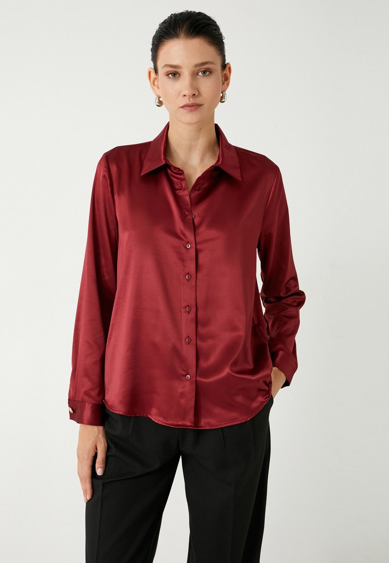 Blusa de satén burdeos con mangas largas, cierre delantero de botones y un cuello clásico. Textura notablemente suave y dobladillo ligeramente curvado.