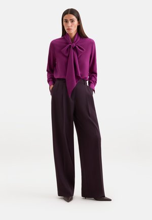 Donna con capelli lunghi e scuri indossa una camicetta viola con un grande fiocco e pantaloni larghi neri, in piedi con le mani nelle tasche su uno sfondo bianco.