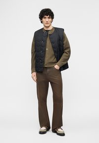 Giovane uomo con capelli ricci che indossa un gilet trapuntato nero sopra un cardigan oliva, pantaloni marroni e mocassini beige con dettaglio a catena, in piedi.