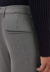Pantalon en laine gris avec une texture lisse, présentant une poche latérale et une coupe ajustée. Associé à un pull à côtes foncé.