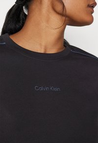 Juodas megztinis su apykakle, turintis mėlyną išsiuvinėtą "Calvin Klein" logotipą ir kontrastinę mėlyną siuvinėjimą. Minkšta, tekstūruota medžiaga.