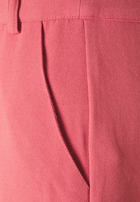 Růžová texturovaná látka s boční kapsou, štíhlým prošíváním a uvolněným střihem. Materiál vypadá měkce a lehce.