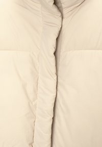 Beige pufferjakke med en glat, skinnende overflade. Har en høj krave, lodrette sømme og en lynlås foran i fuld længde.