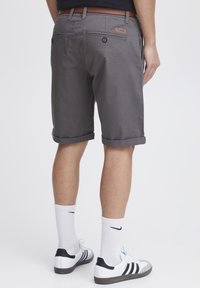 Graue Baumwollshorts mit umgeschlagenen Säumen, ausgestattet mit Gesäßtaschen und einem Leder-Logo-Patch. Getragen mit weißen Socken und schwarz gestreiften Turnschuhen.