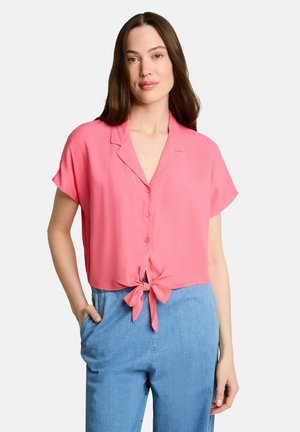 Woman with long brown hair wearing a pink short-sleeve buttoned shirt tied at the waist and light blue high-waist pants, standing with one hand in pocket.