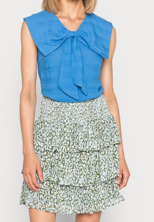 Kobieta ubrana w niebieski, bezrękawnik z dużą kokardą oraz spódnicę o wysokim stanie z wielowarstwowym zielono-białym abstrakcyjnym wzorem.
