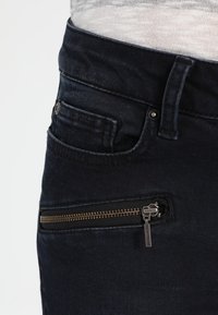 Tmavé džínové kalhoty s postranní kapsou na zip a malým kovovým táhlem, spolu s tradičním designem přední kapsy.