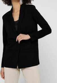 Černý blazer s přizpůsobeným střihem, klopovými reverzy a dvěma předními kapsami. Tkanina má hladkou strukturu; stylizováno přes černý top.