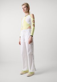 Langarm bodysuit in geel en wit met uitsnijding bij de schouders, gecombineerd met witte wijde broek met zijbranding en groene schoenen.