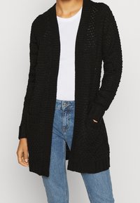 Žena v černém pleteném dlouhém kardiganu přes bílé tričko a modré džíny, s jednou rukou v kapse.