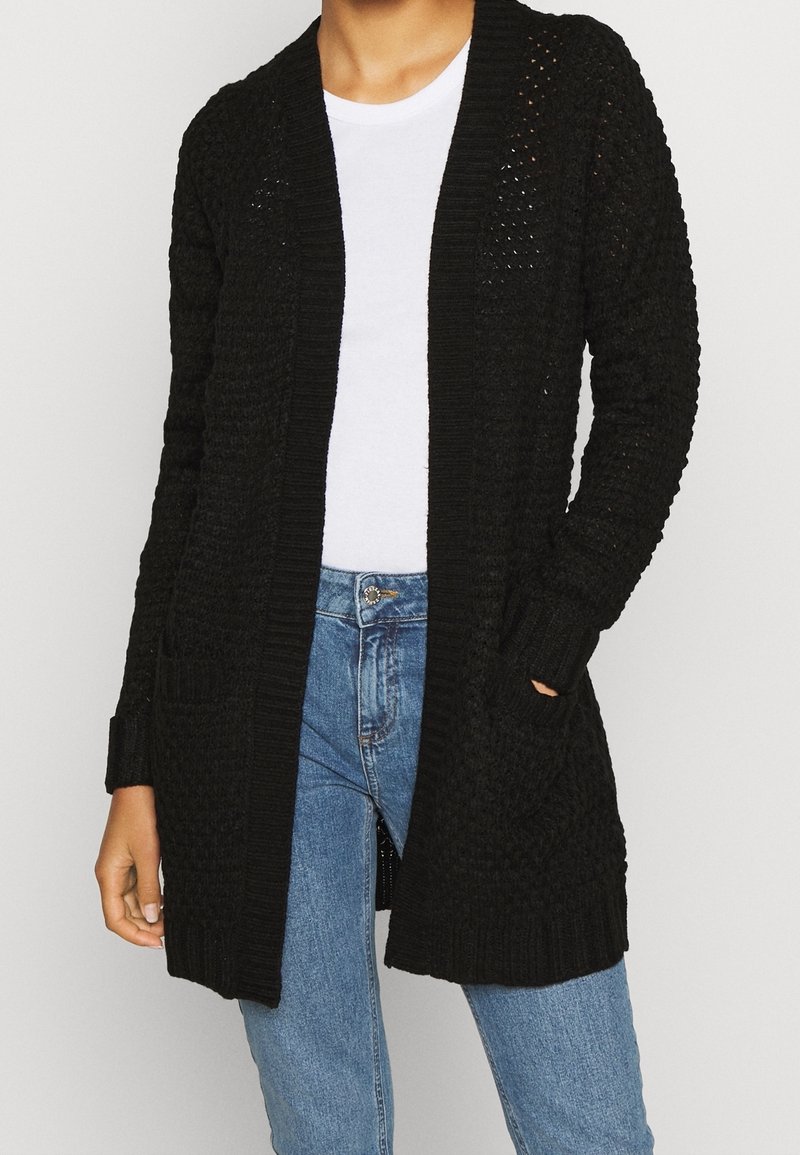 Žena v černém pleteném dlouhém kardiganu přes bílé tričko a modré džíny, s jednou rukou v kapse.