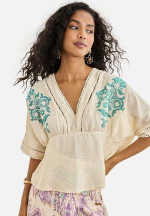 Mujer con cabello rizado que lleva una blusa color crema con bordado floral en tonos verde azulado y mangas sueltas, combinada con pantalones estampados en púrpura y crema.