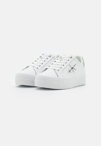 Baskets blanches avec une semelle texturée blanche, dotées d'un pliage lisse et d'accents argentés, marquées "Calvin Klein" sur le côté.