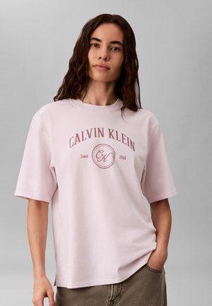 Ung kvinde med mørkt bølget hår iført en lyserød Calvin Klein t-shirt og brune bukser, stående mod en grå baggrund.