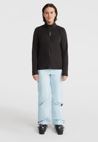 Čierna fleece bunda s vysokým golierom a plným zipsom, skombinovaná s svetlomodrými nepremokavými nohavicami a čiernymi lyžiarskymi topánkami. Jednoduchý športový dizajn.