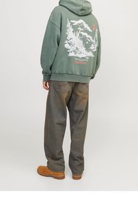 Vihreä huppari, jossa valkoinen graafinen printti takana, yhdistettynä väljiin ruskeisiin farkkuihin ja ruskeisiin saappaisiin. Rentoon tyyliin, ylipitkä istuvuus.